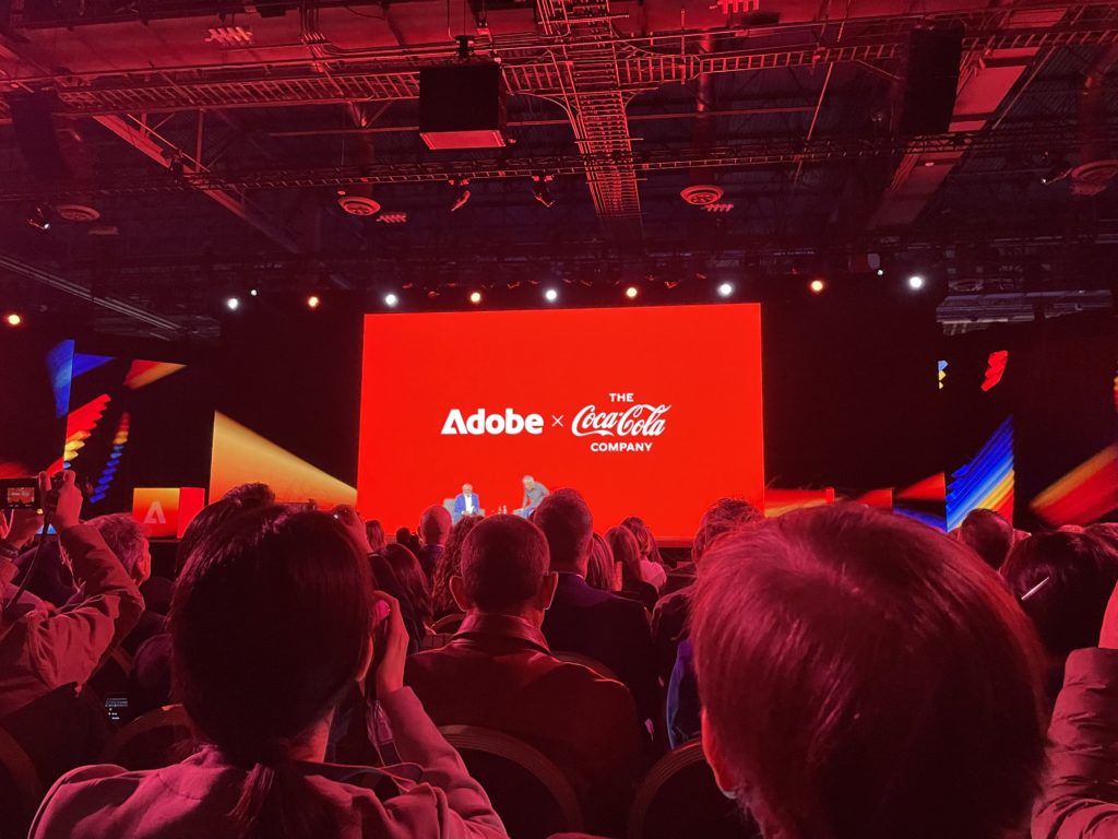 Bühne des Adobe Summit 2025 mit einer großen roten Leinwand, auf der „Adobe x The Coca-Cola Company“ zu sehen ist. Im Vordergrund sitzt das Publikum, einige machen Fotos.